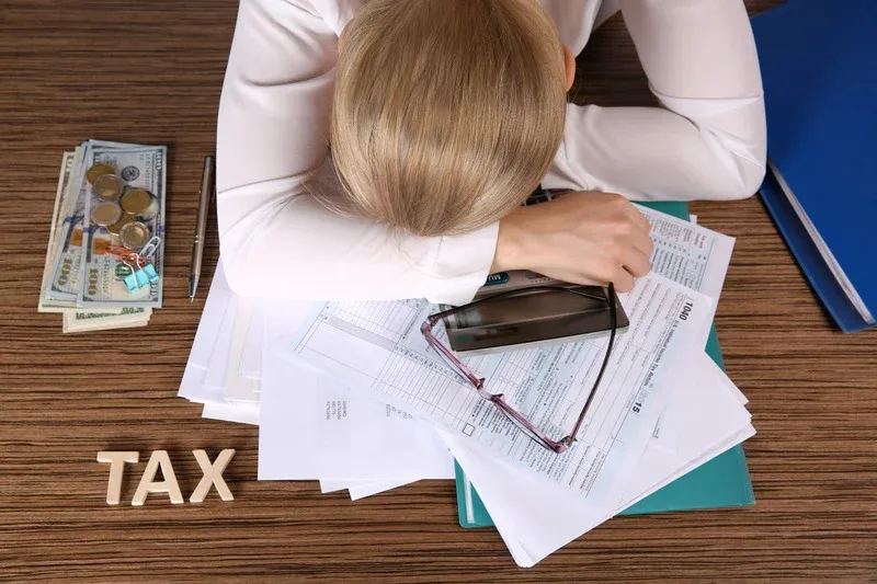 Femme affalée sur un bureau couvert de documents fiscaux, avec un panneau « TAX »